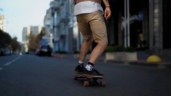 Un uomo intelligente dalla pelle chiara è appassionato di guida veloce su un longboard sulla strada. Le gambe sono in primo piano . — Foto Stock