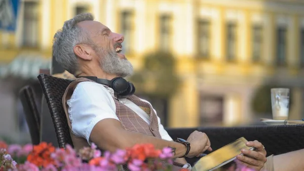 Grey bearded man laughs from the news on a tablet on a sunny day in a cafe — Stock Photo, Image