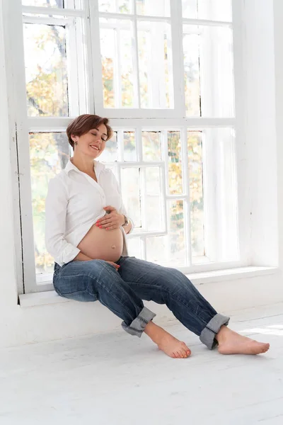 Mulher grávida sorri tocando a barriga no quarto branco — Fotografia de Stock