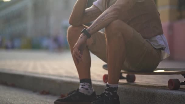 Mann in Lässigkeit kommuniziert auf einem Handy, das auf einem Longboard auf dem Gehweg sitzt — Stockvideo