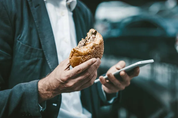 Закрыть портрет молодого белого воротничка, поедающего нездоровую пищу во время работы над смартфоном на открытом воздухе — стоковое фото