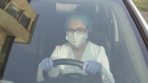 Mujer asiática de mediana edad en una máscara protectora, bata y guantes sostiene el volante del coche — Vídeos de Stock