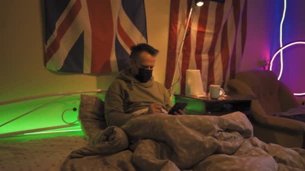 インフルエンザの男は熱い飲み物を飲みながらタブレットでベッドに横たわっている. — ストック動画