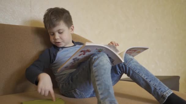 Pojken sitter på soffan läser bok och spelar på smartphone bär jeans. Hemundervisning under karantändagar med föräldrar som arbetar på distans. Prores 422. — Stockvideo