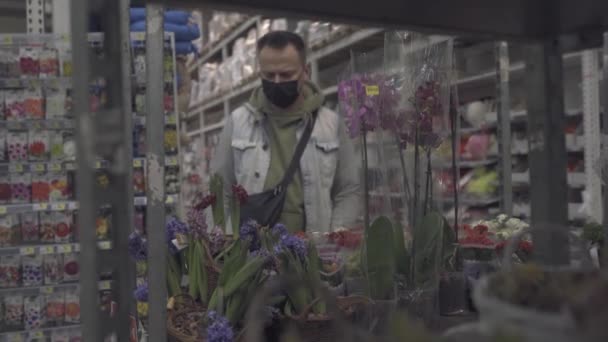 Un acheteur masculin examine les fleurs dans des pots avec un centre commercial. Un homme fait des achats pendant la pandémie de coronavirus dans un masque protecteur et des gants jetables. Il achète une fleur pour sa femme. Prores 422 — Video