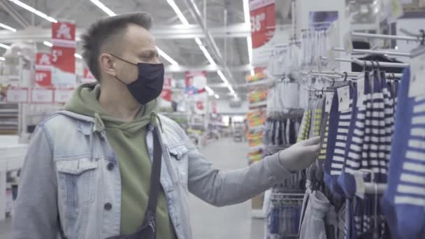 En una máscara protectora en su cara, un joven elige calcetines mientras revisa diferentes opciones en el centro comercial. Compras durante la pandemia del coronavirus. Prores 422. Auchan, Kiev, Ucrania. abril 2020 — Vídeo de stock