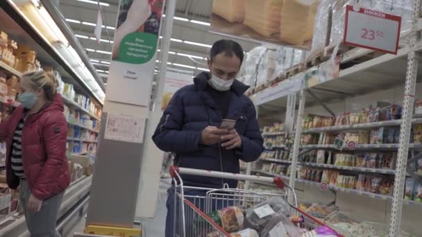 Homem com careca na cabeça escreve uma mensagem para a esposa enquanto faz compras ou responde e-mail de trabalho tardio. Homem com máscara protetora enquanto faz compras em quarentena. Auchan. Kiev, Ucrânia, abril de 2020 Prores 422 — Vídeo de Stock