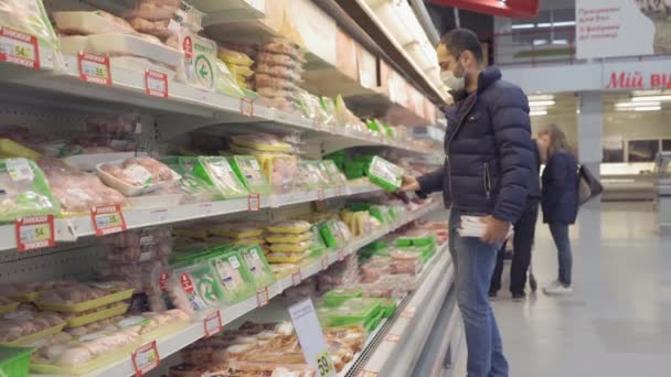 Mavi ceketli ve kot pantolonlu genç bir adam marketten paketlenmiş et seçiyor. Karantinada alışveriş yaparken koruyucu maskeli bir adam. Auchan. Kiev, Ukrayna, Nisan 2020. Prores 422 — Stok video