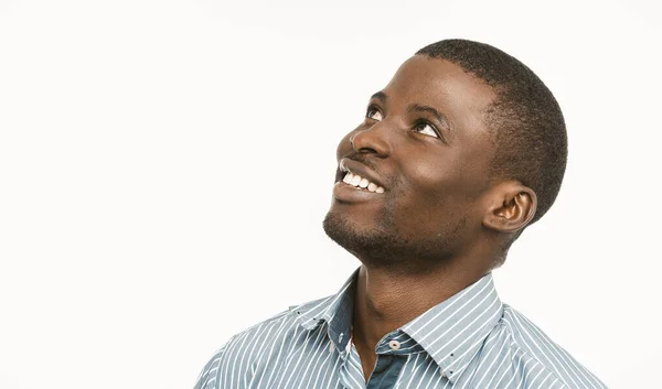 Afro-américain bel homme toothy sourit en levant les yeux et sur le côté, jeune homme à la peau foncée en chemise à rayures bleues isolé sur fond blanc. Espace de copie situé sur le côté gauche. Image tonique — Photo