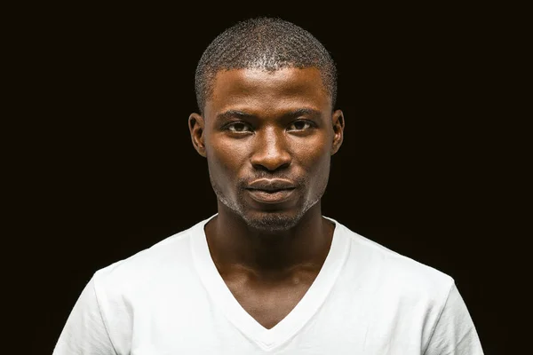 Serious African american man looking at the camera, attractive dark-skinned young guy in a white t-shirt posing expressing confidence isolated on black background. Toned image — Stock Photo, Image