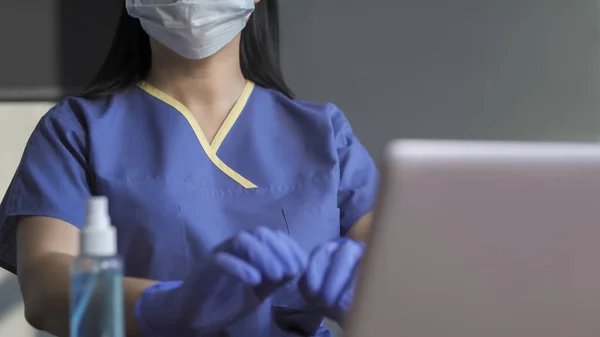 Medic fêmea em uniforme azul funciona no computador sentado à mesa com desinfetante sobre ele. Copiar espaço no lado direito. Fechar tiro — Fotografia de Stock