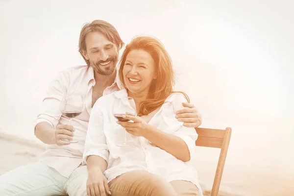 Glückliches Paar verbringt Zeit zusammen in der Nähe des Meeres. Mann und Frau trinken Rotwein, lächeln und umarmen sich, während sie draußen am Wasser sitzen. Getöntes Bild — Stockfoto