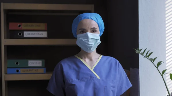 Femeia doctor stând în biroul ei. Femeia serioasă în mască de protecție și uniformă albastră se uită la cameră în timp ce stă pe fundalul dosarelor cu înregistrări medicale — Fotografie, imagine de stoc