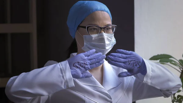 Стомлений жіночий лікар у білій формі та захисній масці показує, що напис зупиняє вірус на його долонях у гумових рукавицях, дивлячись збоку. Пандемія коронавірусу — стокове фото