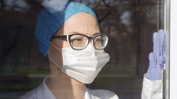 Väsynyt lääkäri katsoo ikkunaa Touch Glass käsin. Naispuolinen lääkintämies odotti pandemian loppua. Lääketieteen ja terveydenhuollon käsite — kuvapankkivalokuva