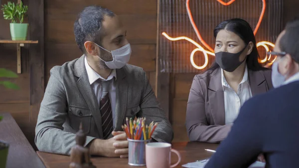 Team of business people are busy with teamwork during coronavirus outbreak. Colleagues wearing protective masks hold a business meeting while sitting at the negotiating table in modern office — Stock Photo, Image