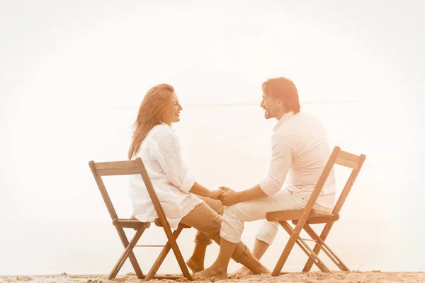 Mature couple resting at beach holding hands. Happy man and woman holding hands and looking each other while sitting on sandy beach on sea background. Sunset concept. Toned image — Stock Photo, Image