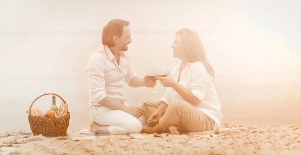Romantisches Picknick am Meer. Mann und Frau unterhalten sich beim Rotweintrinken. Reifes Paar sitzt auf Sand und schaut einander zu. Obstkorb mit einer Flasche Wein daneben. Getöntes Bild — Stockfoto