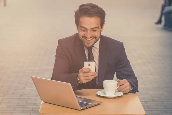 Бізнесмен використовує смартфон, що працює на відкритому повітрі. Щасливий чоловік дивиться на мобільний телефон, сидячи за столом з комп'ютером і чашкою кави на ньому на вуличному кафе. Бізнес-концепція. Вид спереду. Тоноване зображення — стокове фото