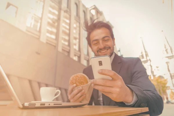 Бізнесмен їсть гамбургер за допомогою телефону на вулиці. Усміхнена людина в костюмі їжі в кафе швидкого харчування на вулиці сучасного європейського міста. Тонограма — стокове фото