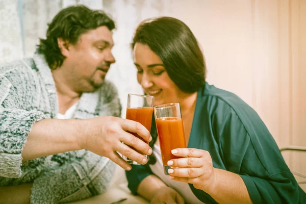 笑超重夫妇结合眼镜与橙汁。男人在厨房桌子边说了句节日祝酒词 — 图库照片