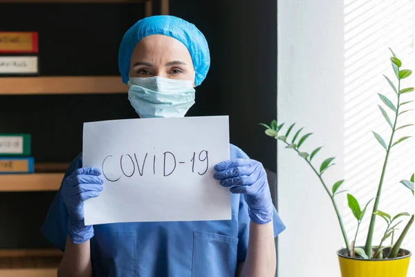 Enfermeira ou médico de uniforme azul segurando uma folha de papel com inscrição covid-19. Uma jovem médica está no escritório a olhar para a câmara. Conceito de surto de coronavírus — Fotografia de Stock