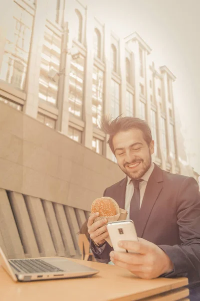 Businessman using phone eating sandwich or hamburger outdoors. Happy man in suit sits in fast food cafeteria on the city street. Toned image. Vertical format — Stock Photo, Image