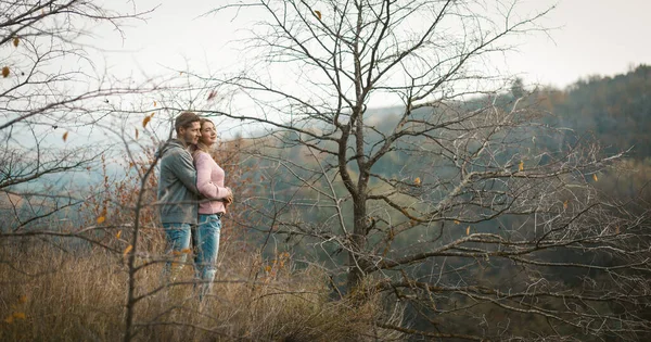 Abrazando pareja en el amor se encuentra en una ladera mirando hacia abajo, hombre joven y una mujer admiran el bosque de otoño que crece en las laderas de abajo —  Fotos de Stock