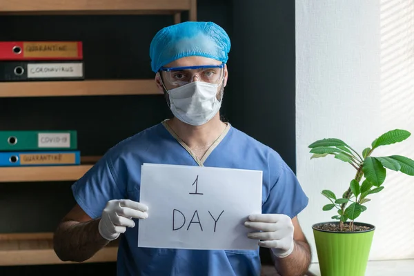 Médico cansado en uniforme protector anuncia un día antes del final de la pandemia de coronavirus. Médico masculino sostiene una hoja de papel con la inscripción un día. Concepto de cuenta regresiva —  Fotos de Stock