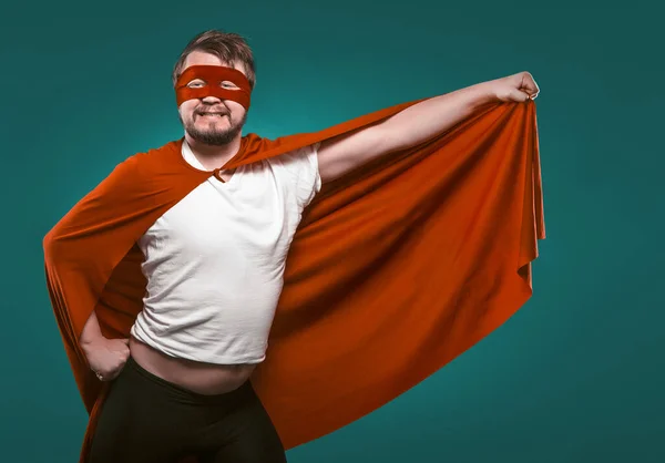 Hombre divertido super héroe listo para volar salvar el mundo. Hombre sonriente con máscara y traje de superhéroe rojo sosteniendo su capa en su mano posando sobre fondo verde vizcaíno — Foto de Stock