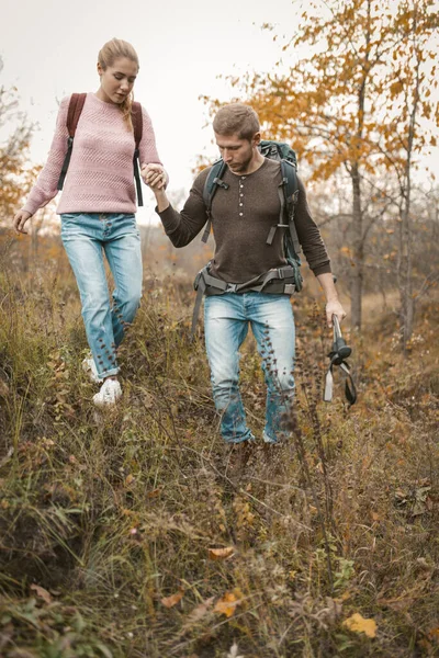 El hombre está apoyando a una hermosa rubia con una mochila caminando juntos en la naturaleza salvaje en otoño — Foto de Stock