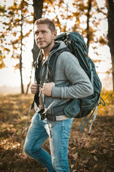 Backpacker reist in de natuur. Serieuze blanke man met een grote rugzak kijkt in de verte terwijl hij tegen de achtergrond van een herfstbos staat — Stockfoto
