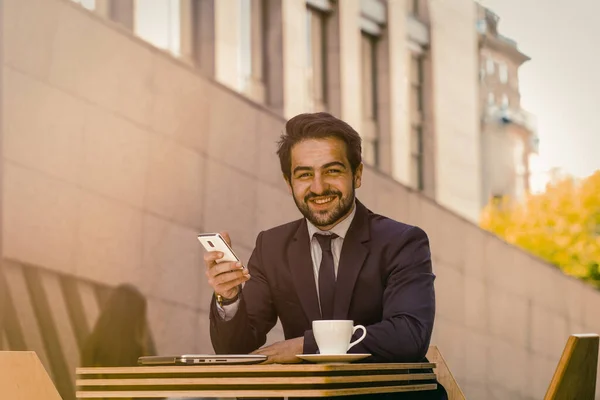 Бізнесмен використовує мобільний телефон на відкритому повітрі. Успішний чоловік зубний посміхається тримаючи смартфон, сидячи за столом з чашкою кави на ньому в міському вуличному кафе. Бізнес на концепції ходу. Тоноване зображення — стокове фото