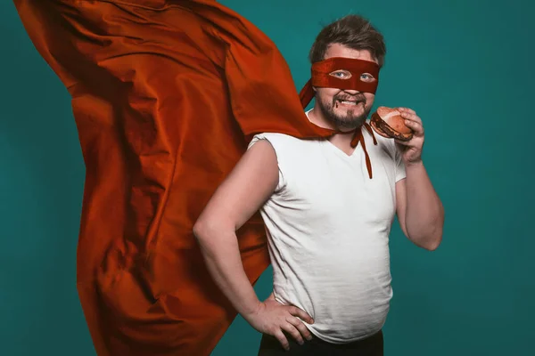 Az éhes szuperhős ember hamburgert eszik, szuperhőst vagy antihőst piros ruhában, repülő köpenyben, és gyorskaját eszik, miközben a kamerába néz. Vágd ki a zöld háttér — Stock Fotó