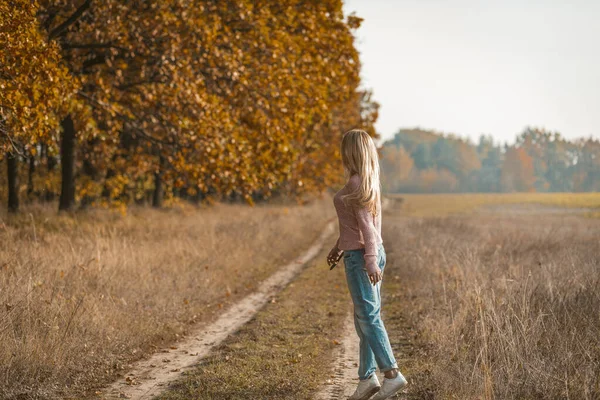 Vista trasera de la mujer joven en ropa casual de puntillas en la naturaleza, encantadora joven rubia caminando por un camino de tierra cerca del bosque de otoño —  Fotos de Stock