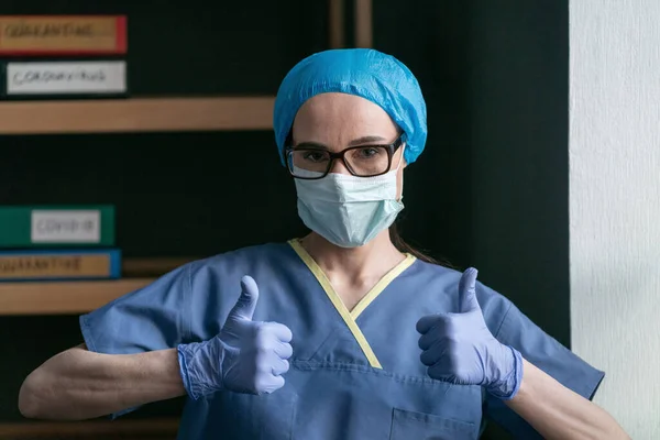 Asistenta arată degetele în sus semn oferind prognostic pozitiv. Femeia medic în uniformă de protecție albastră și mănuși de unică folosință arată degetele mari gestul în picioare în birou — Fotografie, imagine de stoc