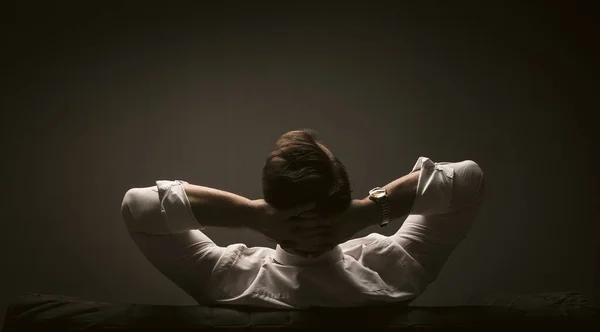 Rear view of Businessman thinking while sitting at couch. Man in white shirt resting with hands behind his head on dark grey background — Stock Photo, Image