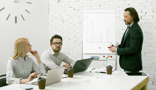 Aantrekkelijke zakenman staat in de buurt van een wit bord presenteert het team zijn zakelijke idee. Collega 's luisteren aandachtig naar de spreker die aan de kantoortafel zit. Teamwork in office concept — Stockfoto