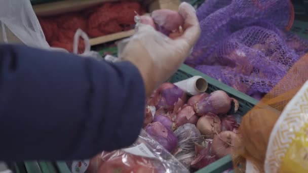 Een man sorteert uien in een winkel. Winkelen tijdens de quarantaine periode, een man in een wegwerp plastic handschoen selecteert een boog uit een groot aantal op het product display. Prores 422 — Stockvideo