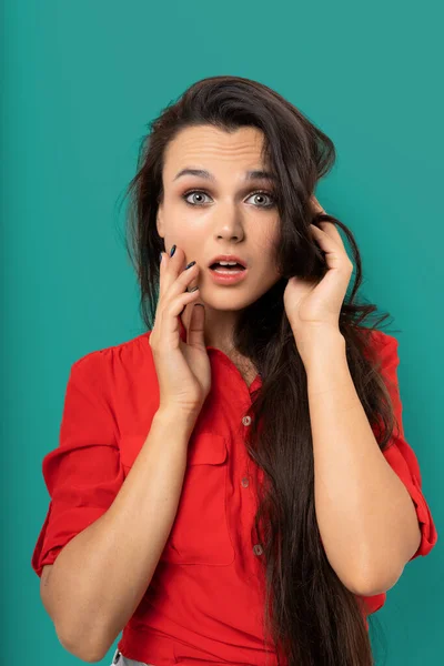 Mujer sorprendida toca su cara con las manos, Mujer bastante mestiza con la boca abierta y el pelo largo y oscuro en blusa roja aislada en Vizcaya Fondo Verde. Concepto de venta. Concepto de emoción —  Fotos de Stock