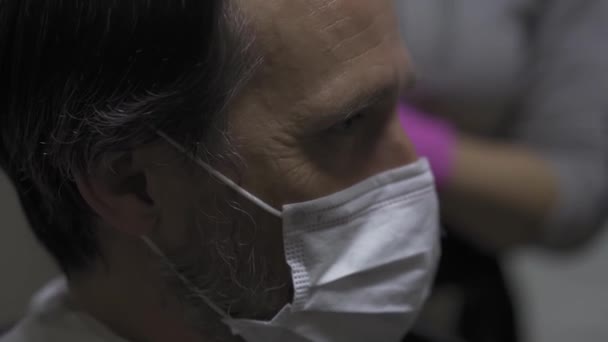 Ένας άντρας με προστατευτική μάσκα κάθεται στο κομμωτήριο σε ένα κούρεμα, ο κομμωτής προετοιμάζει το κεφάλι για χτένισμα. Καραντίνα κουρέων. Πρότυπα 422 — Αρχείο Βίντεο