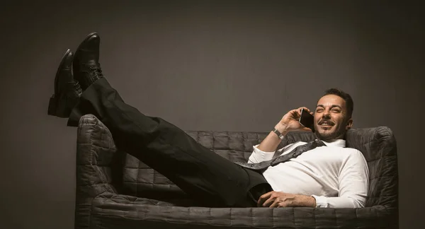 Empresário a comunicar através do telemóvel. Sorrindo homem bonito falando em um telefone inteligente enquanto deitado em um sofá e jogou suas pernas em calças pretas e botas de couro no sofá — Fotografia de Stock