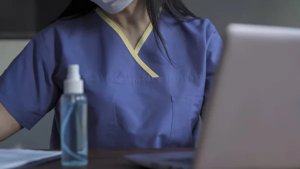 Fáradt orvos dolgozó számítógép. Védőmaszkos nő és egyenruhás kitöltő papír nyomtatvány, miközben laptoppal dolgozik az orvosi rendelőben a világjárvány idején. Közelkép. — Stock Fotó