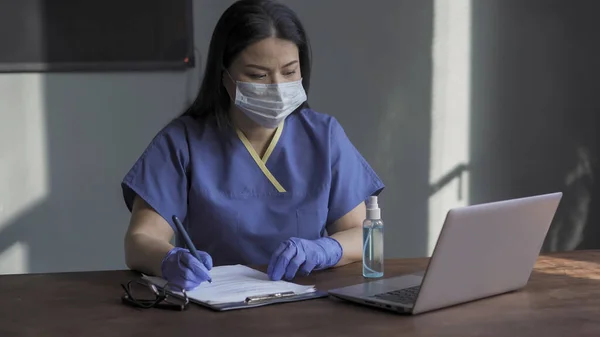 Médica feminina preenche formulário de papel enquanto trabalha com laptop em consultório médico. Mulher em máscara protetora e uniforme funciona durante uma pandemia — Fotografia de Stock