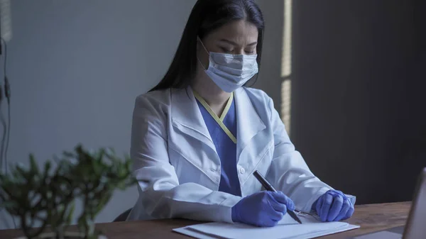 El doctor cansado escribe en papel en blanco sentado en la mesa. Médico femenino en uniforme protector que trabaja en su oficina — Foto de Stock