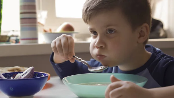 Голодный ребенок дошкольного возраста ест суп из миски. Серьезный мальчик забил полный рот супа, сидя с большими щеками. Хорошая концепция аппетита — стоковое фото