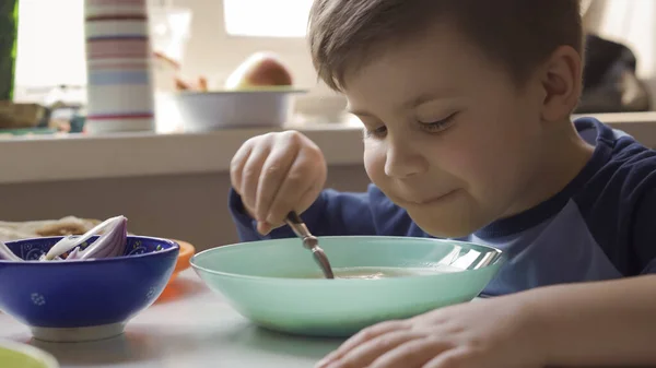 Маленький мальчик ест суп один, сидя за обеденным столом. Мальчик-дошкольник остается дома в дни карантина или изоляции. Портрет крупного плана — стоковое фото