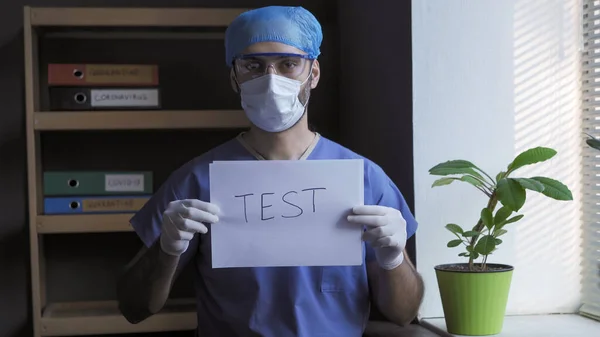 Medicul se oferă să facă un test medical. Omul în uniformă de protecție albastră stă aproape de fereastră uitându-se la cameră în timp ce ține testul de slogan pe foaia de hârtie. Conceptul de pandemie de coronavirus — Fotografie, imagine de stoc