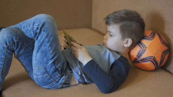 Preescolar mira la pantalla del teléfono móvil tumbado en el sofá con la cabeza descansando en la pelota de fútbol. Niño estudiando en línea debido a la cuarentena. Concepto de educación en el hogar. Vista del perfil — Foto de Stock