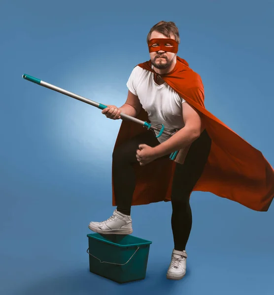 Superbohater gotowy do sprzątania. Człowiek w masce i czerwonym płaszczu superbohatera pozujący z wiadrem i mopem przedstawiającym się jako muzyk rockowy. Odizolowany na niebieskim tle — Zdjęcie stockowe